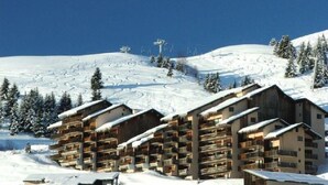A snow-covered mountain with a ski lift in the background and a row of wooden buildings with balconies in the foreground.