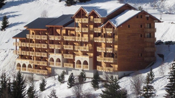 Un grand chalet en bois doté de nombreux balcons et de fenêtres en arc de cercle, entouré de montagnes et d’arbres couverts de neige.