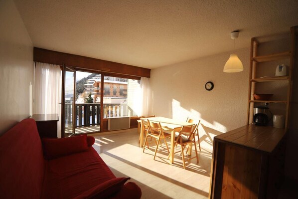A room with a red sofa, a wooden dining table with chairs, a kitchen area with a coffee maker, and a balcony with a view of buildings.
