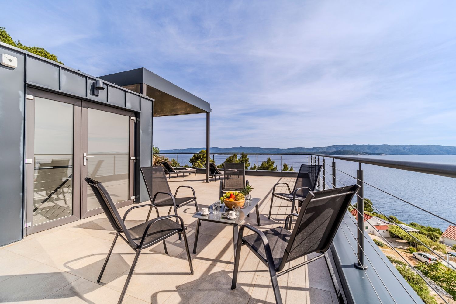 A modern outdoor patio with a table and chairs, overlooking a body of water and mountains.
