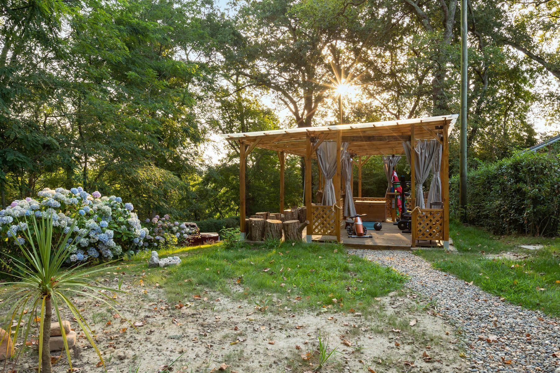 Un kiosque en bois doté d’un plancher ajouré, entouré d’un jardin où poussent des hortensias et un palmier.