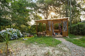 Ein hölzerner Pavillon mit Gitterboden, umgeben von einem Garten mit Hortensien und einer Palme.
