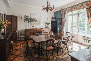 Ein Esszimmer mit einem großen Holztisch, Stühlen, einer Kandelaber und Blick ins Freie durch ein mit Vorhängen drapiertes Fenster.