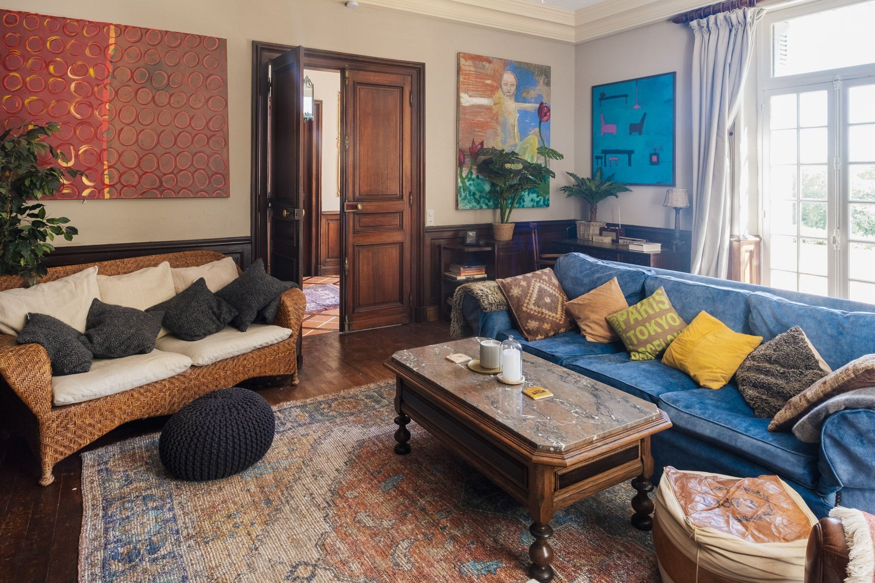 Een woonkamer met een rieten bank, een blauwe bank met gestreepte kussens, een houten salontafel en een tapijt met een patroon.