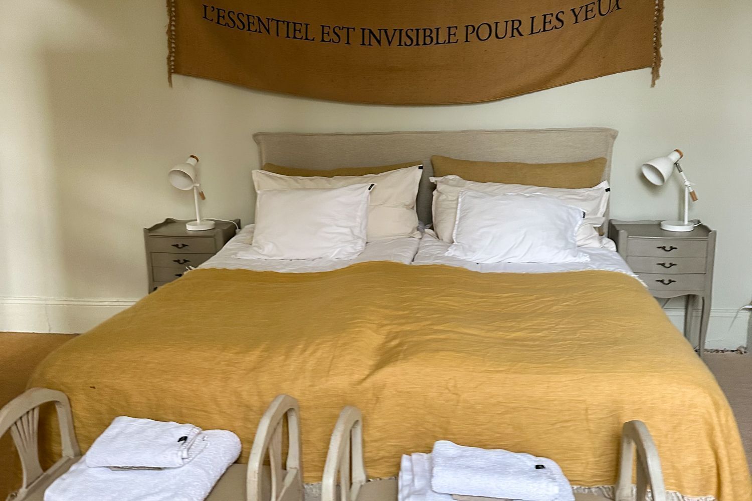 A neatly made bed with a mustard-colored blanket, white pillows, and a headboard. There are two bedside tables with lamps, and a sign above the bed.