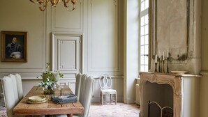 A dining room with a wooden table, white upholstered chairs, a chandelier, a fireplace, and a framed portrait on the wall.