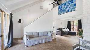Un vaste espace de vie comprenant un canapé de couleur claire, un autre canapé plus foncé et une table basse. On y trouve également un ventilateur de plafond et un grand tableau abstrait accroché au mur.