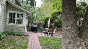 Un patio trasero con un sendero de ladrillo, una parrilla cubierta, mobiliario exterior y un árbol.