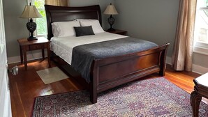 Un dormitorio con una cama grande, dos mesitas de noche, una mesilla auxiliar, un tapete estampado y una ventana con cortinas.