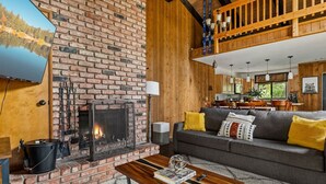 A cozy living room with a brick fireplace, a sofa with yellow cushions, and a television mounted on a brick wall. - Pet Friendly + Ski In Out Unobstructed Views (Big Bear Lake)