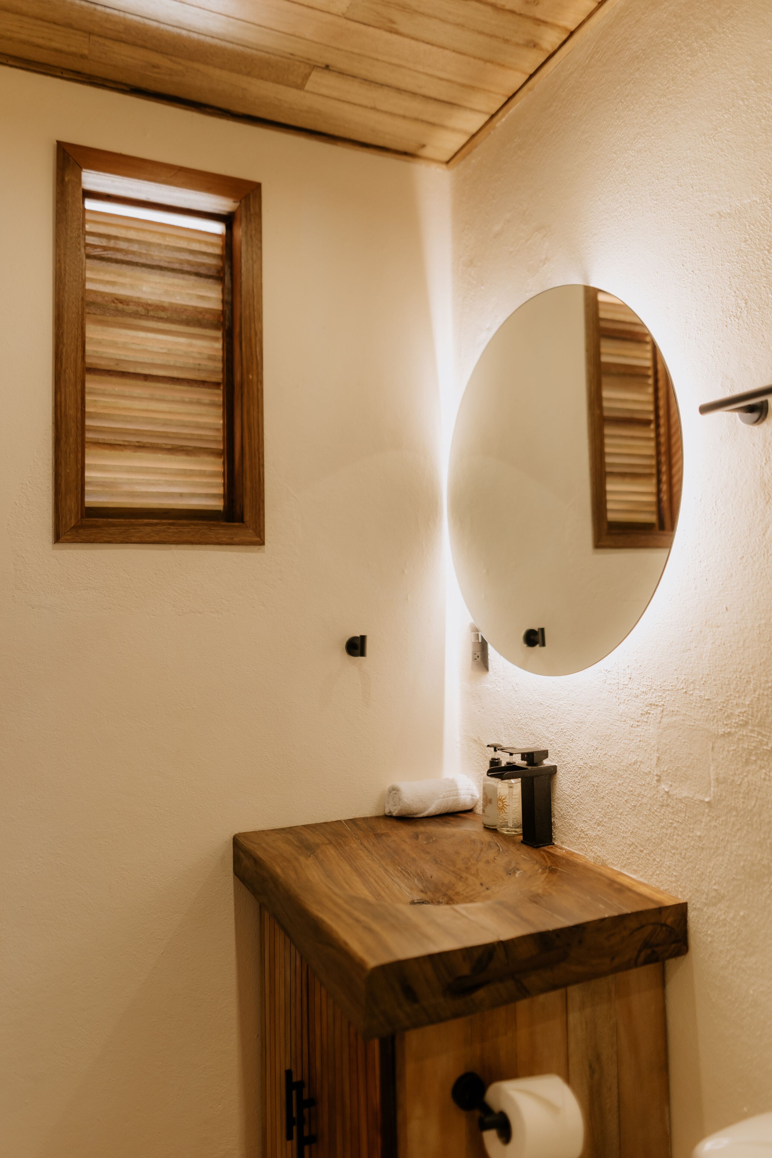 A bathroom with a wooden vanity, a round mirror, and a window with blinds.