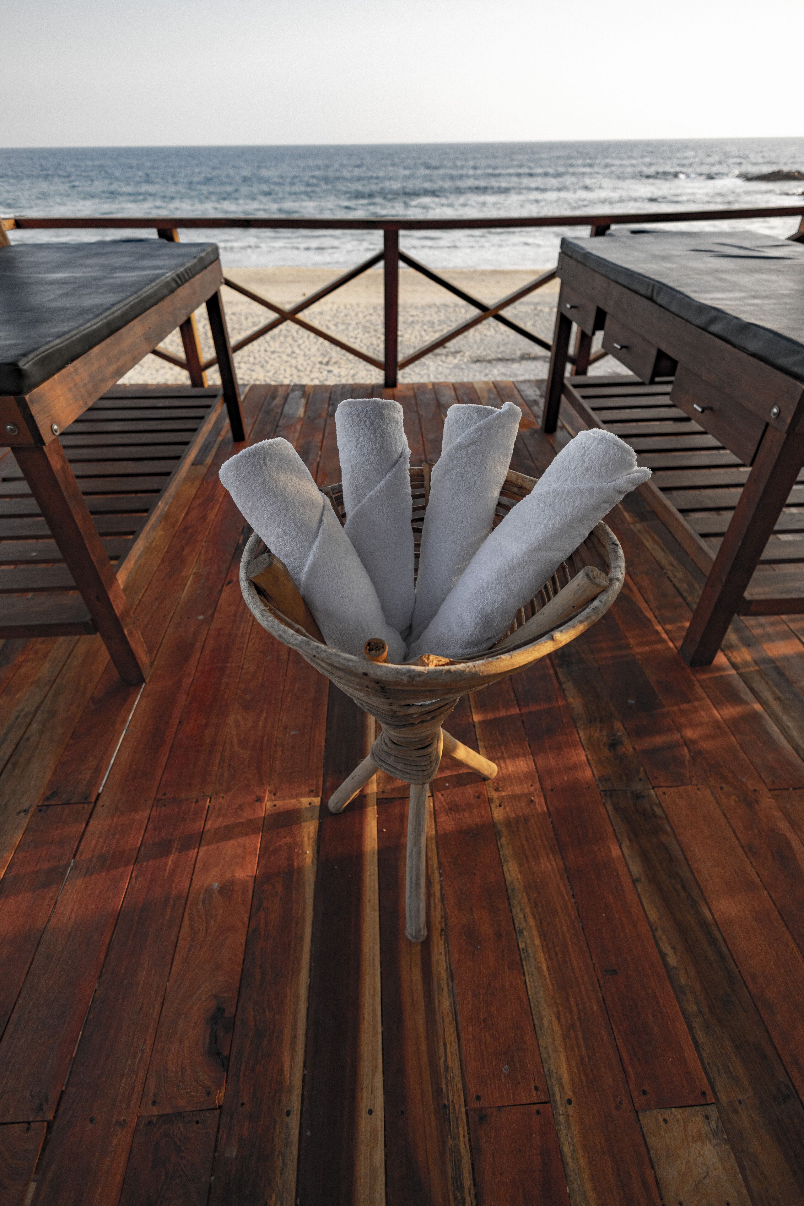 A beachside wooden deck with a woven basket holding white towels, overlooking the ocean.