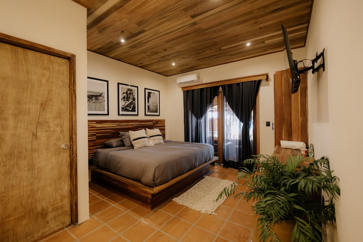 Un dormitorio con techo de madera, una cama con cabecera, un ventanal grande con cortinas y un televisor montado en la pared.