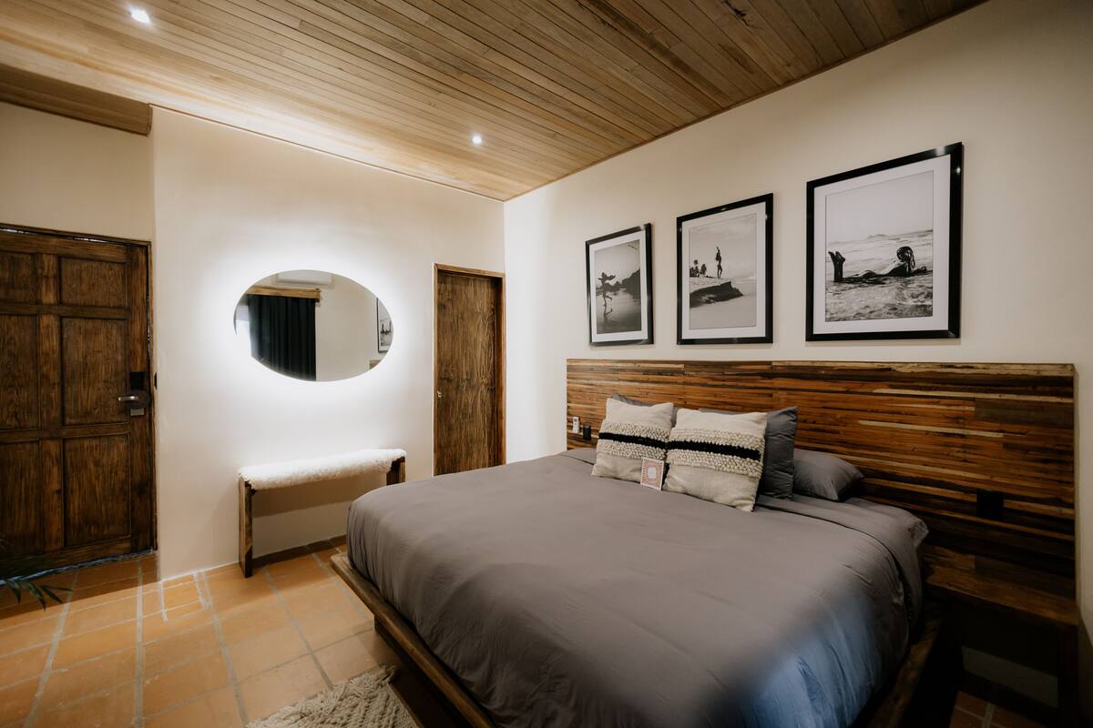 Un dormitorio con cabecera de madera, una cama grande, un banco, un espejo redondo y tres cuadros en la pared.