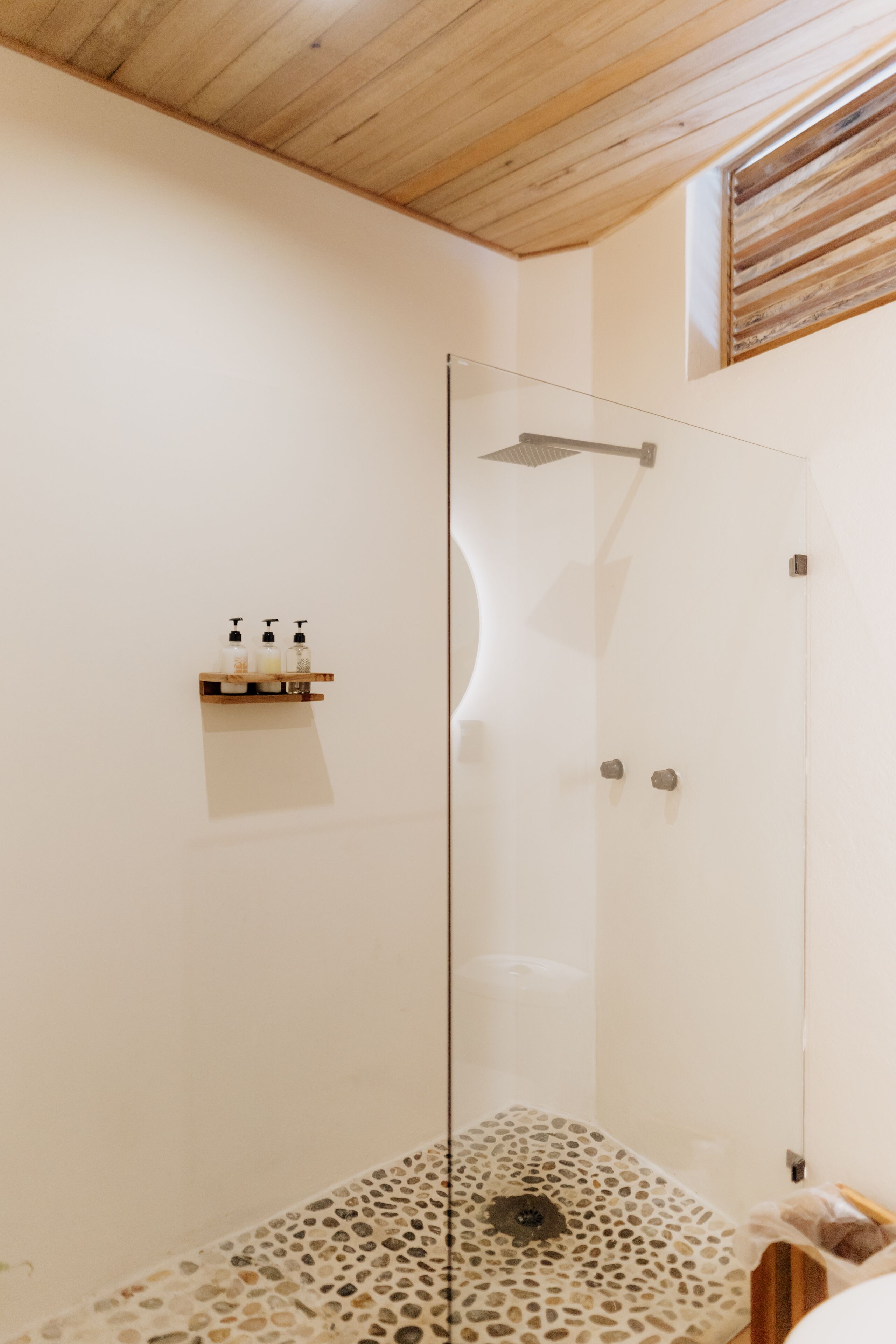 Un espace douche clos par des parois de verre, avec un plafond en bois, une petite étagère contenant des articles de toilette et un sol à motifs.