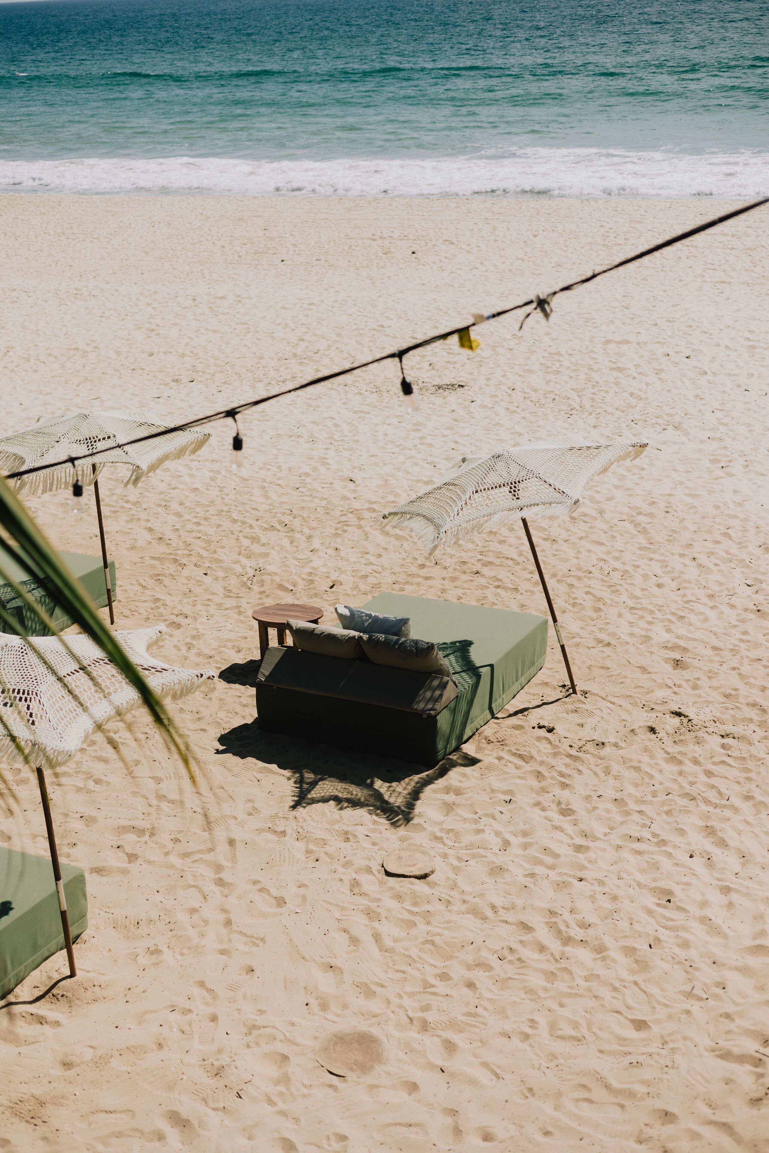 Una playa con una única silla de descanso verde, una pequeña mesa, luces colgantes y un paraguas de playa.