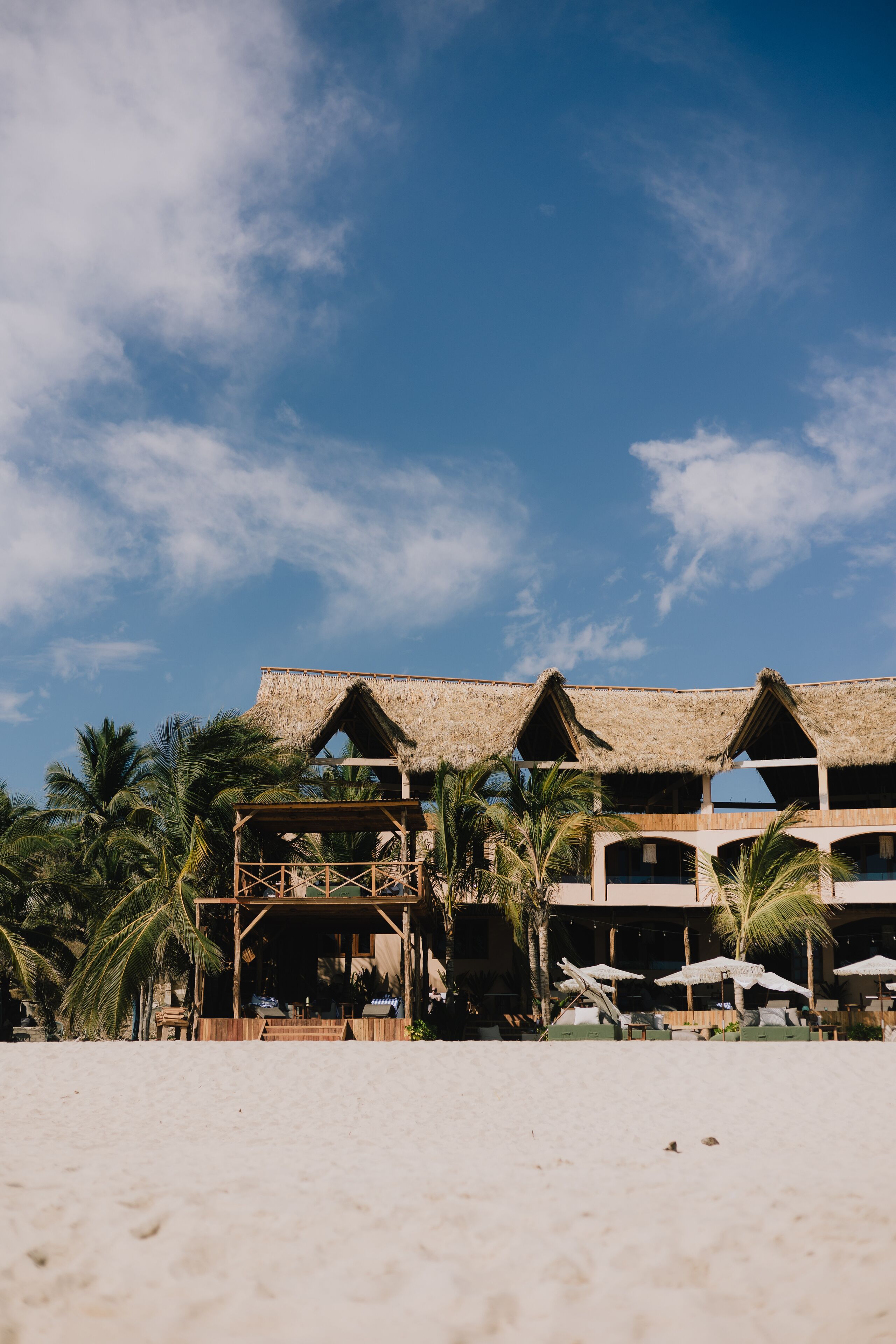 Um hotel à beira-mar com construções de telhado de palha, palmeiras e um céu azul límpido.