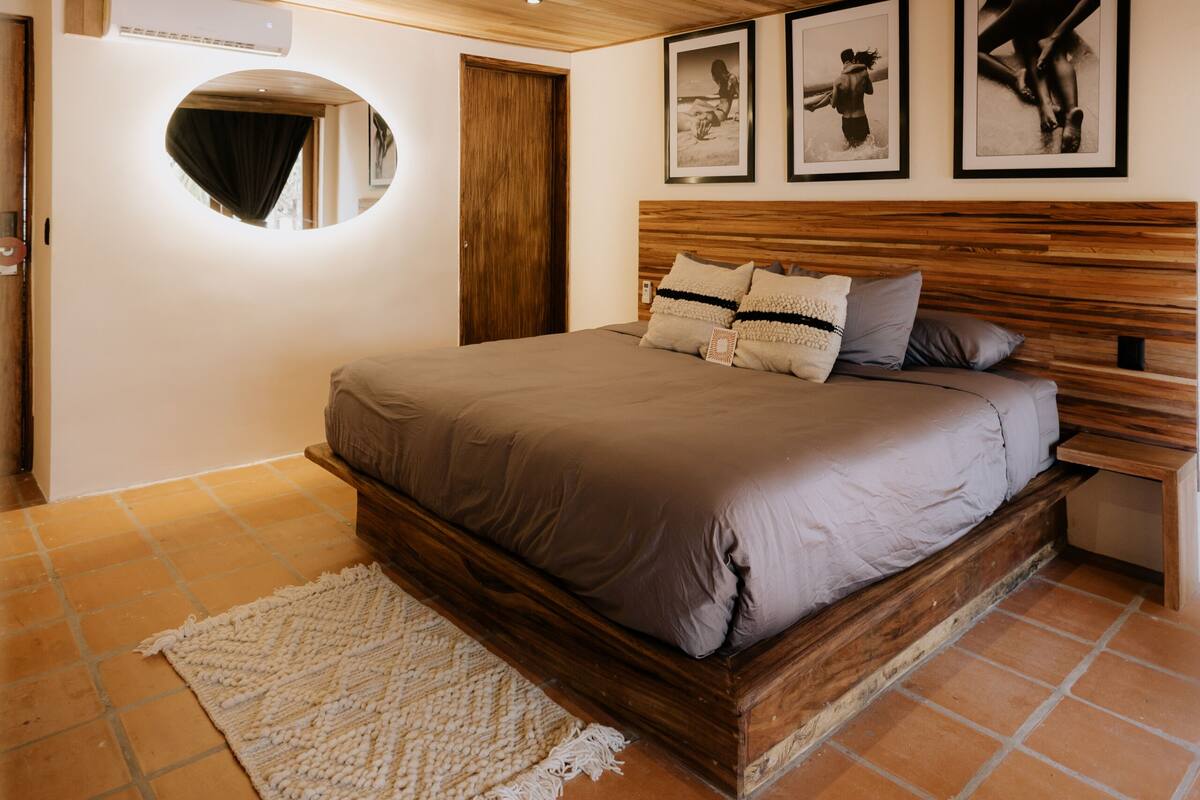 Un dormitorio con techo de madera, una cama grande con ropa de cama gris, un espejo redondo y tres cuadros en la pared.