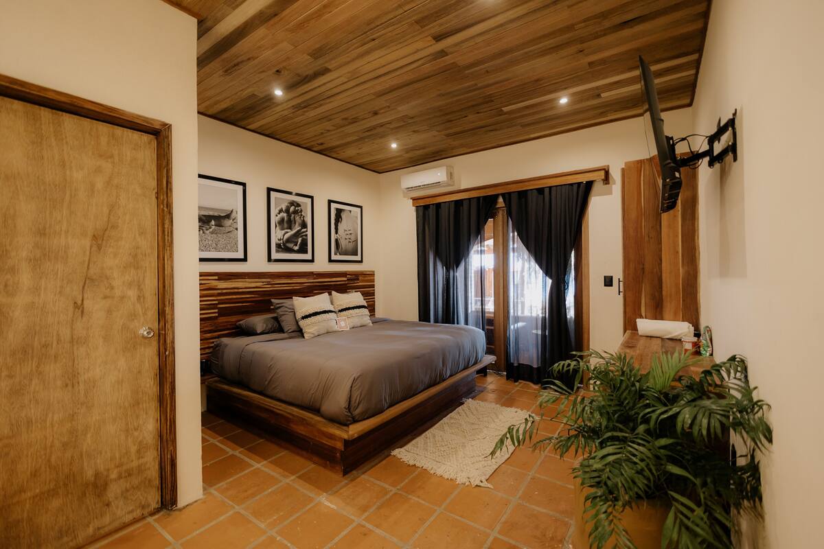 Un dormitorio con techo de madera, una cama con cabecera, un televisor fijado a la pared y una ventana con cortinas.