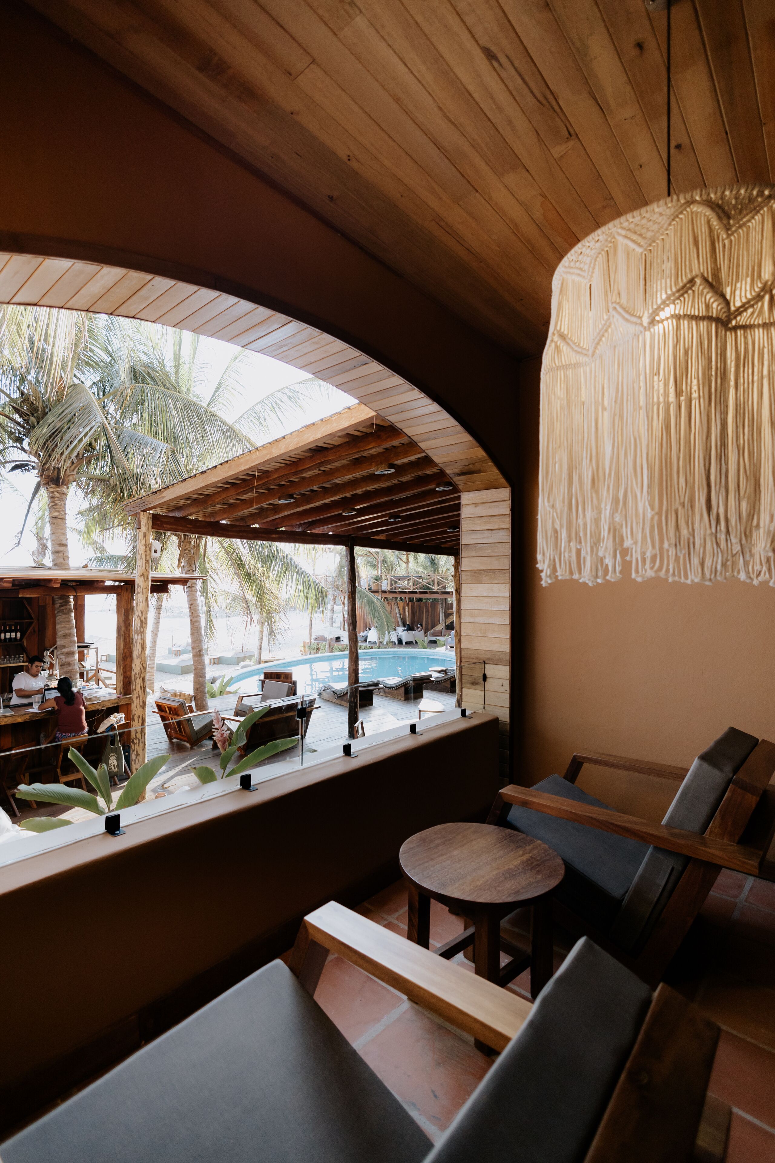 Interior de madeira com vista para uma piscina e palmeiras, com áreas de estar e um lustre pendurado.