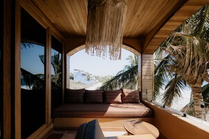 Techo de madera con lámpara colgante, banca de madera con cojines y vista a palmeras y edificios.