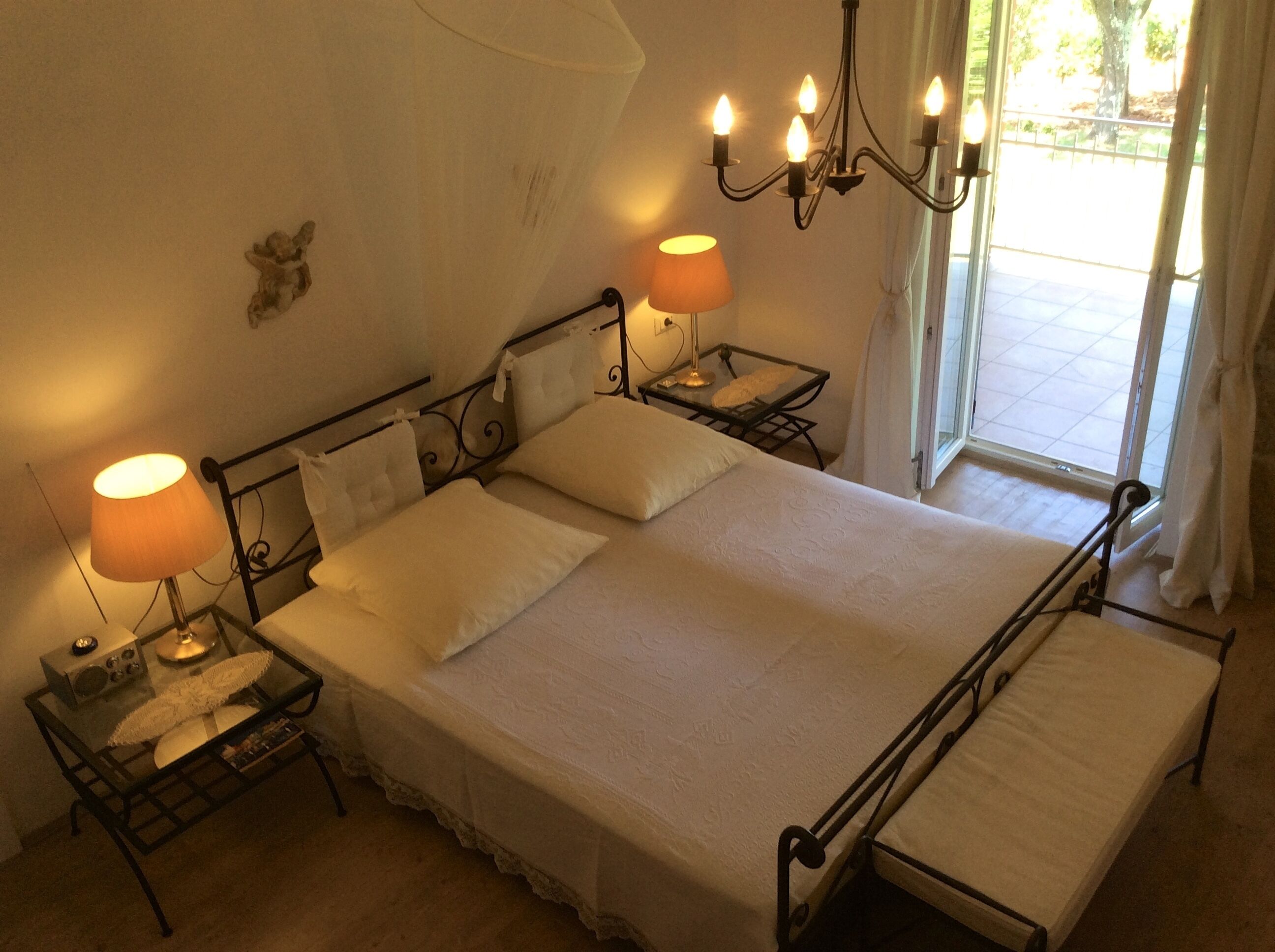 Un letto rifatto con due cuscini e un comodino con una lampada. Sopra il letto c'è un lampadario e una finestra con tende.