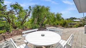 A round white table with benches, surrounded by a garden with trees and shrubs.