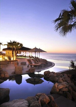A resort pool with lounge chairs and umbrellas, surrounded by rocks and palm trees, overlooking the ocean. - LUXURY STUDIO 5🎖️ LOS CABOS, SAN LUCAS MEX.  (Cabo San Lucas)