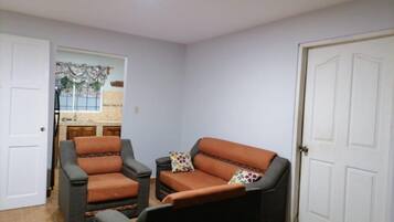 A room with two armchairs and a sofa, a kitchen area visible through an open door, and a white door on the right.