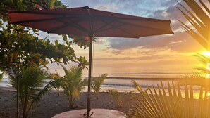 Een tafel aan het strand met een parasol, omringd door palmbomen en een uitzicht op de oceaan bij zonsondergang.