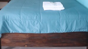 Een net opgemaakt bed met een blauwgroene spreidek en kussens, een houten hoofdbord, een klein nachtkastje en een airconditioningunit aan de muur.