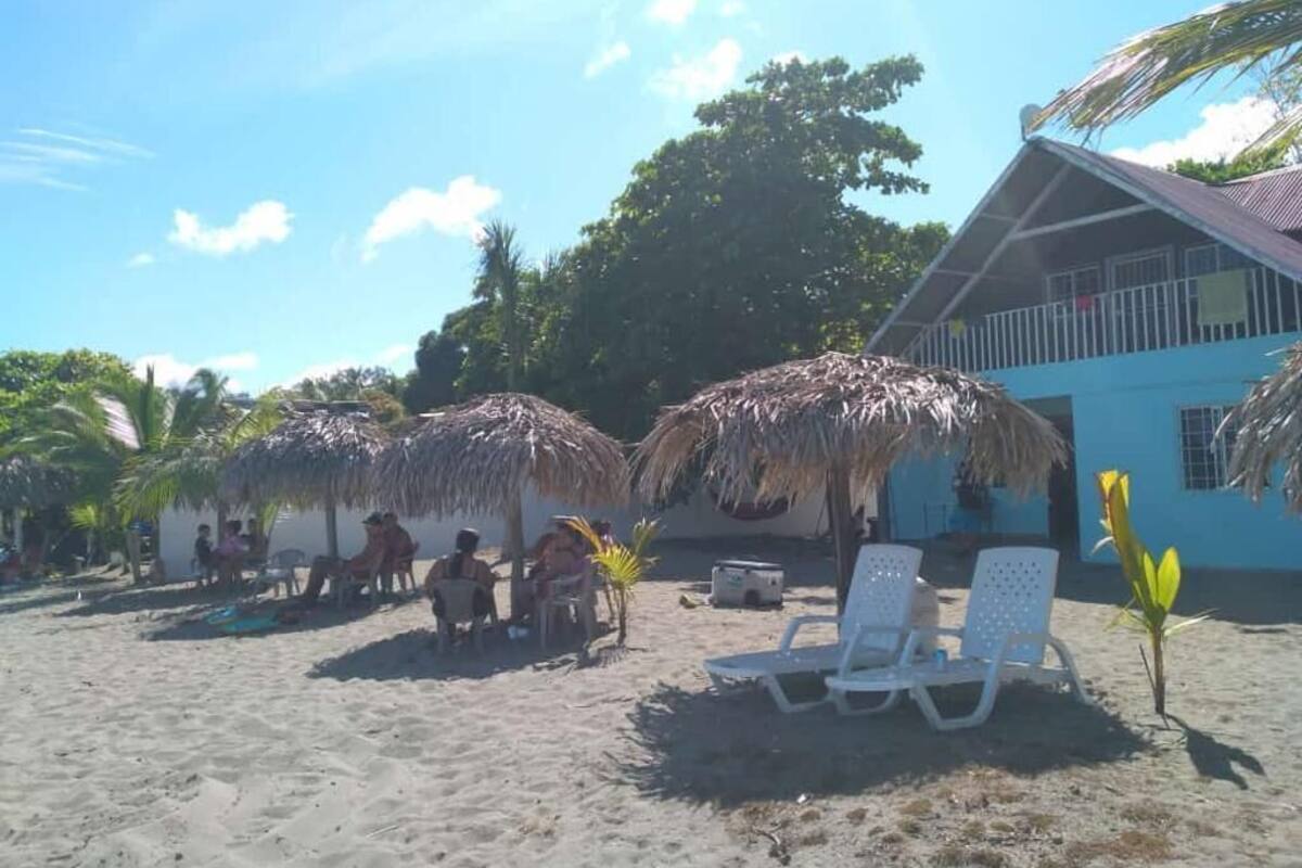 Un cadre idyllique au bord de la plage, avec des parasols en chaume, des chaises longues et un bâtiment à la façade bleue.