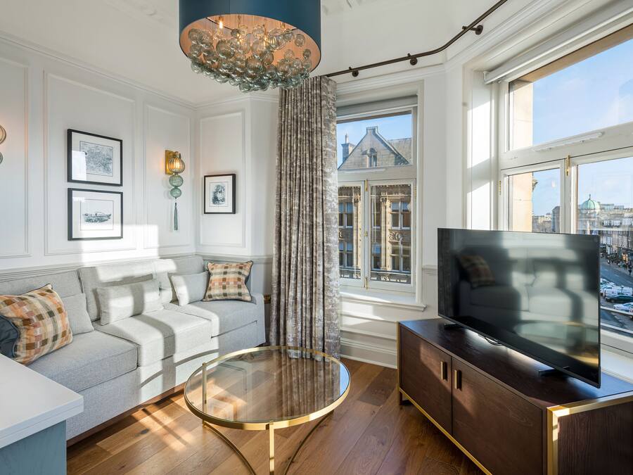 Una sala de estar moderna con un sofá, una mesa de centro de vidrio, un televisor y vistas a un paisaje urbano a través de amplios ventanales.