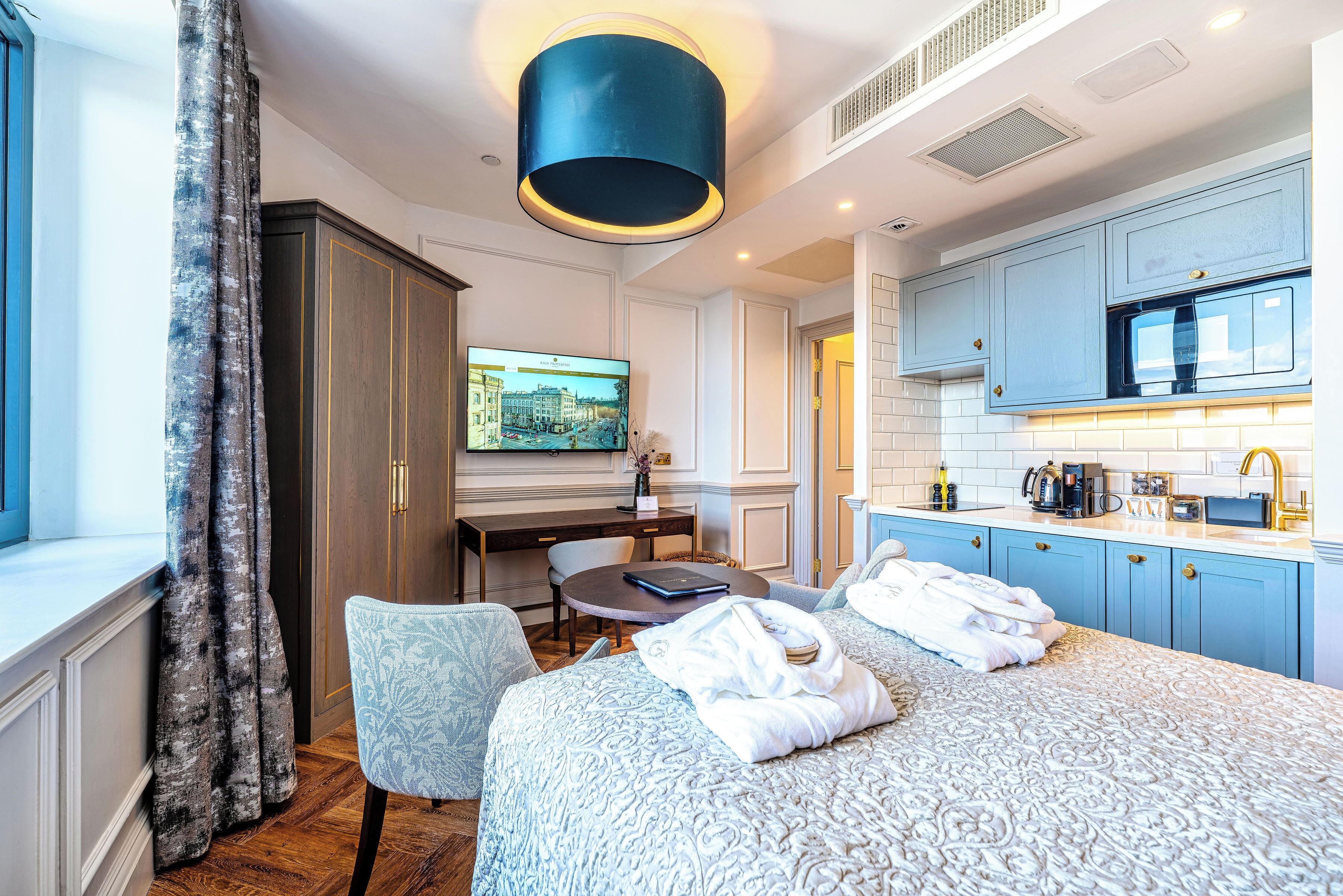 A hotel room with a bed, a desk, a chair, a window with curtains, and a kitchenette.