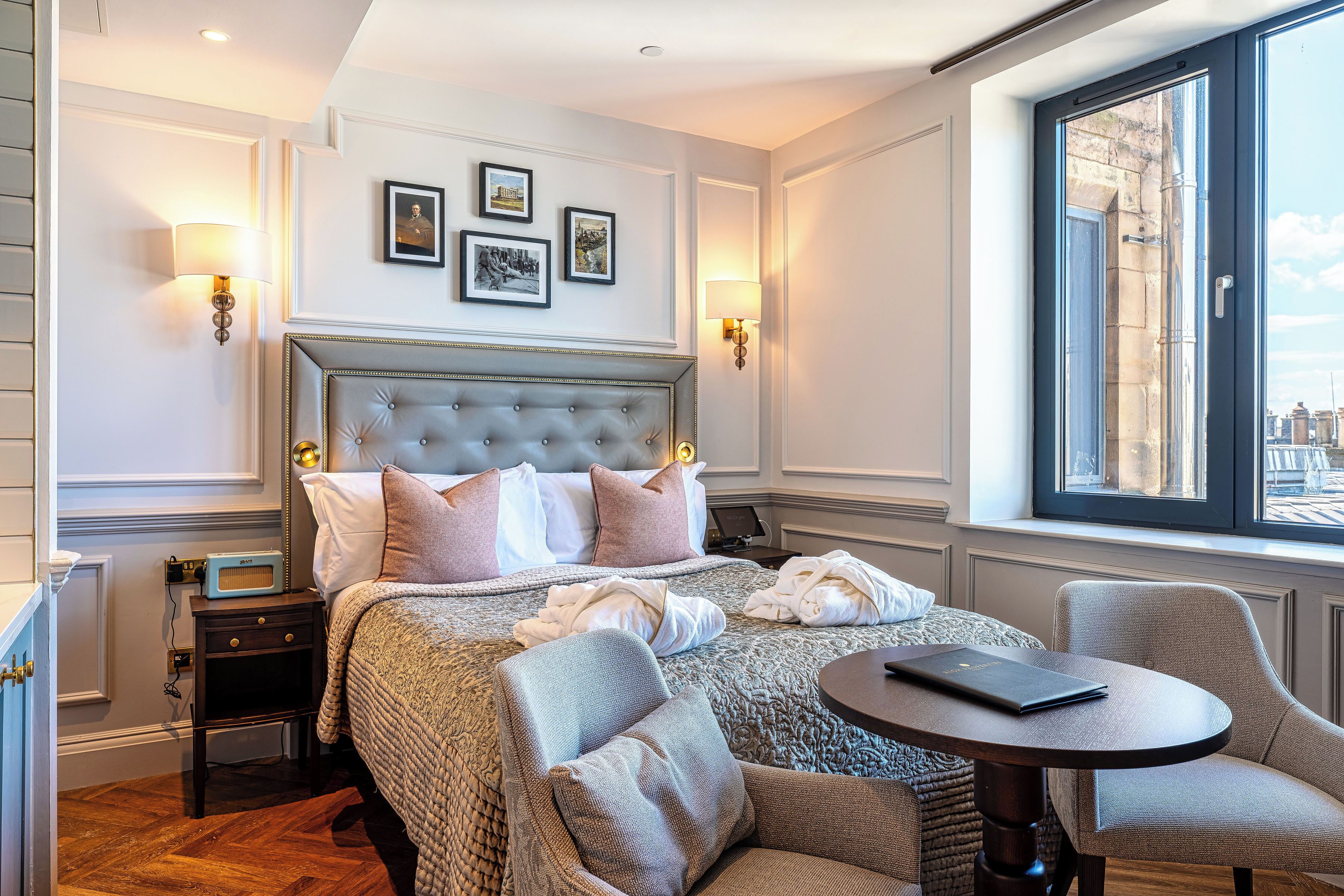 Quarto de hotel com uma cama grande, uma mesa redonda e vista para a cidade através de uma janela ampla.