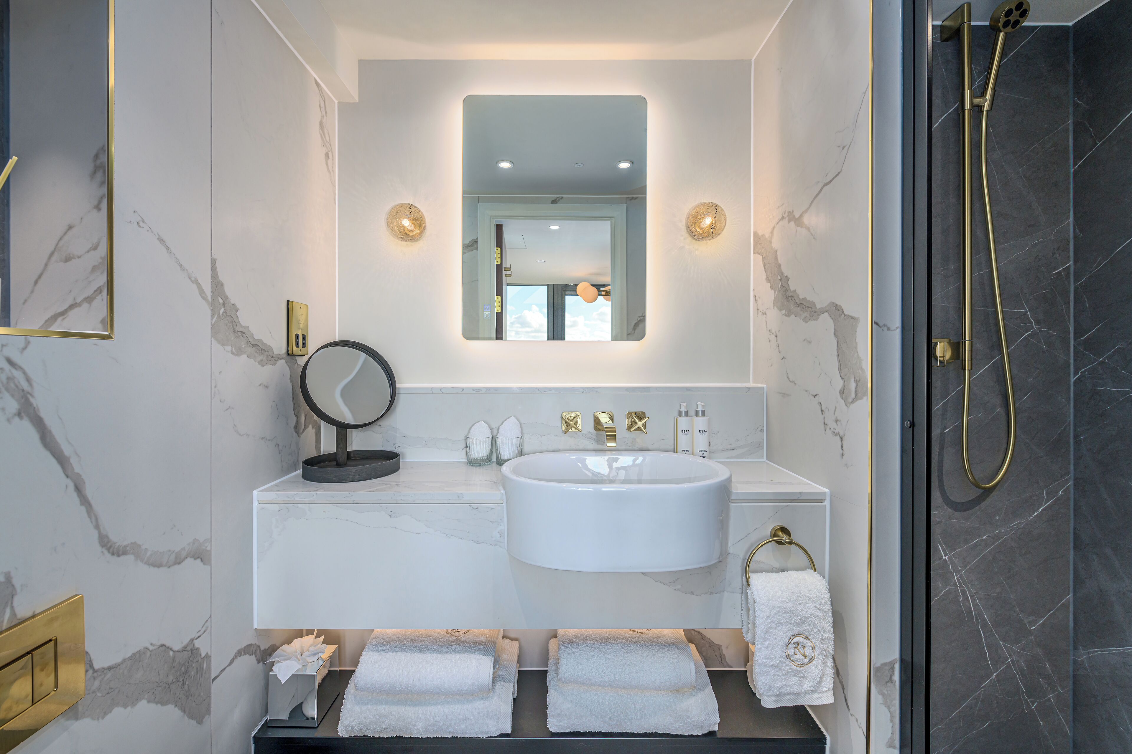 A modern bathroom with a marble sink, a large mirror, and a shower with a gold fixture.