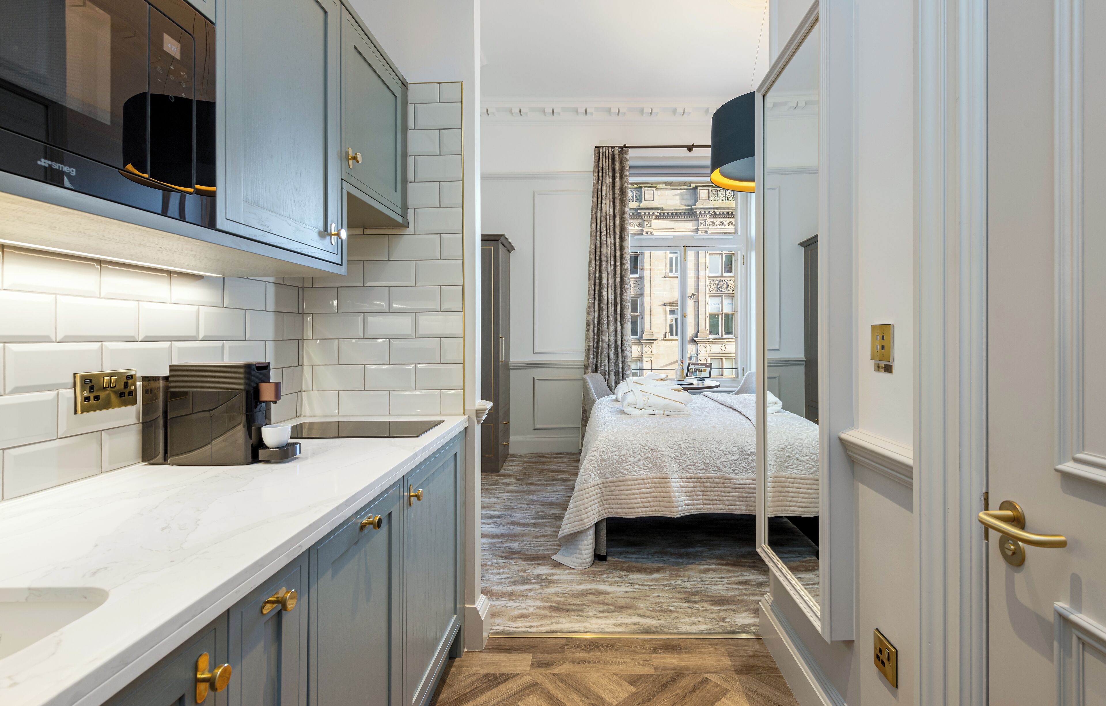 A narrow hallway with a kitchen on one side and a bedroom on the other, featuring a bed with a white duvet, a wooden floor, and a view of a building through a window with curtains.