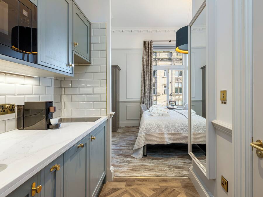A narrow hallway with a kitchen on one side and a bedroom on the other, featuring a bed with a white duvet, a wooden floor, and a view of a building through a window with curtains.