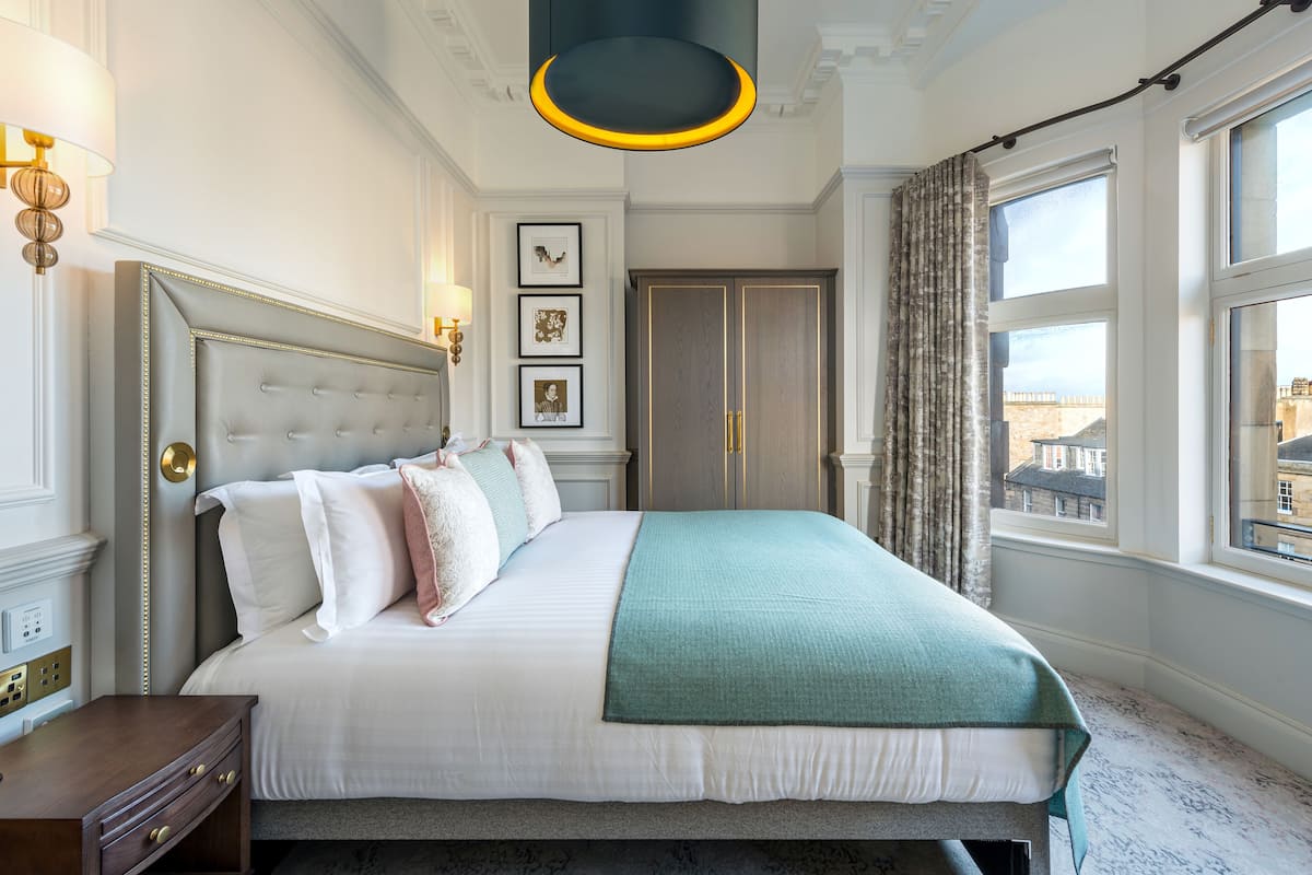 Un lit bien fait, recouvert d’une couvre-lit blanche et turquoise, avec une table de chevet en bois et une lampe fixée au mur.