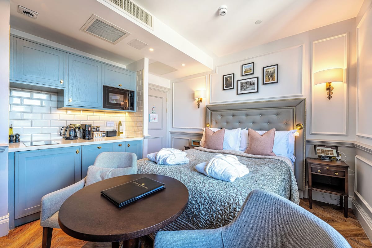 Une chambre d’hôtel moderne dotée d’une kitchenette, d’un grand lit avec plusieurs oreillers, d’une table de chevet avec un livre et d’une petite table de chevet avec une lampe.