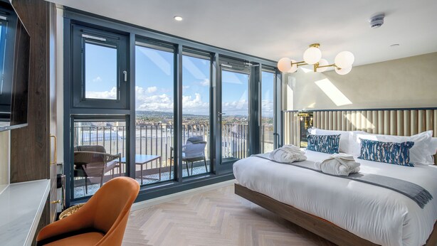 Une chambre d’hôtel moderne dotée d’un grand lit, d’un coin salon avec un fauteuil et d’un balcon offrant une vue sur la ville.