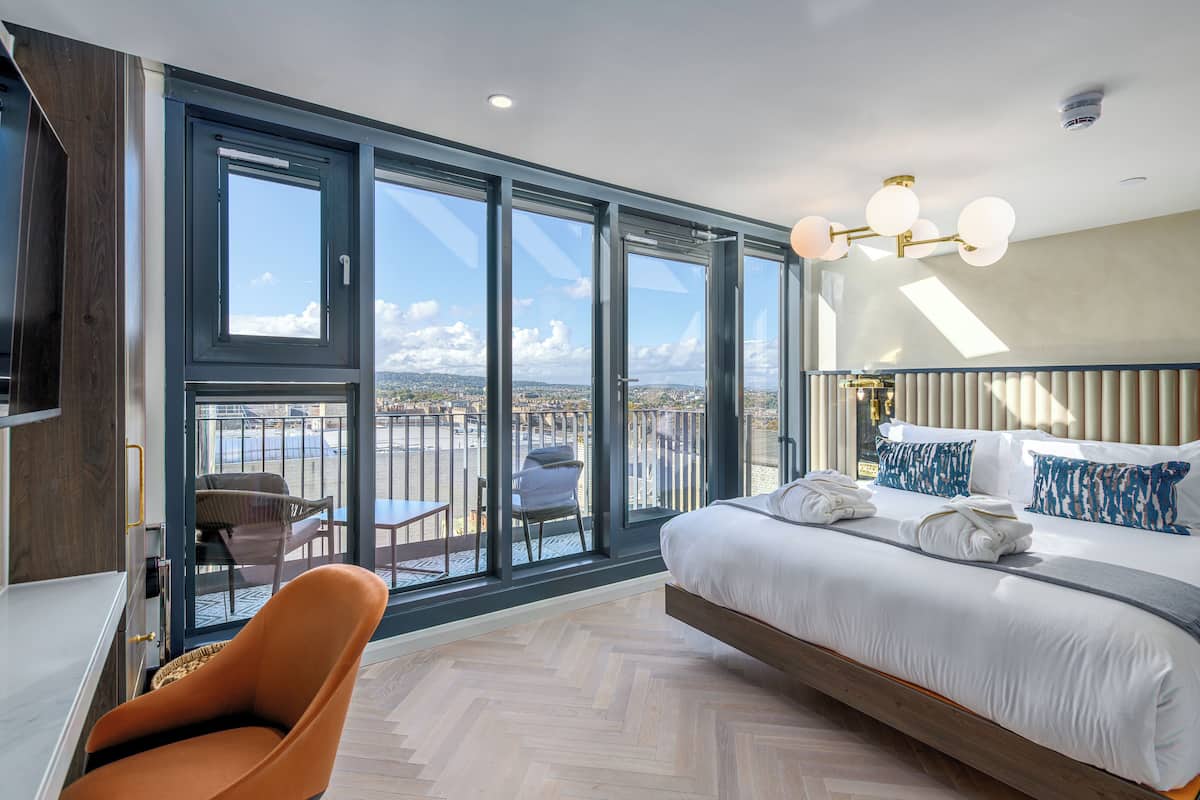 Une chambre d’hôtel moderne dotée d’un grand lit, d’un coin salon avec un fauteuil et d’un balcon offrant une vue sur la ville.