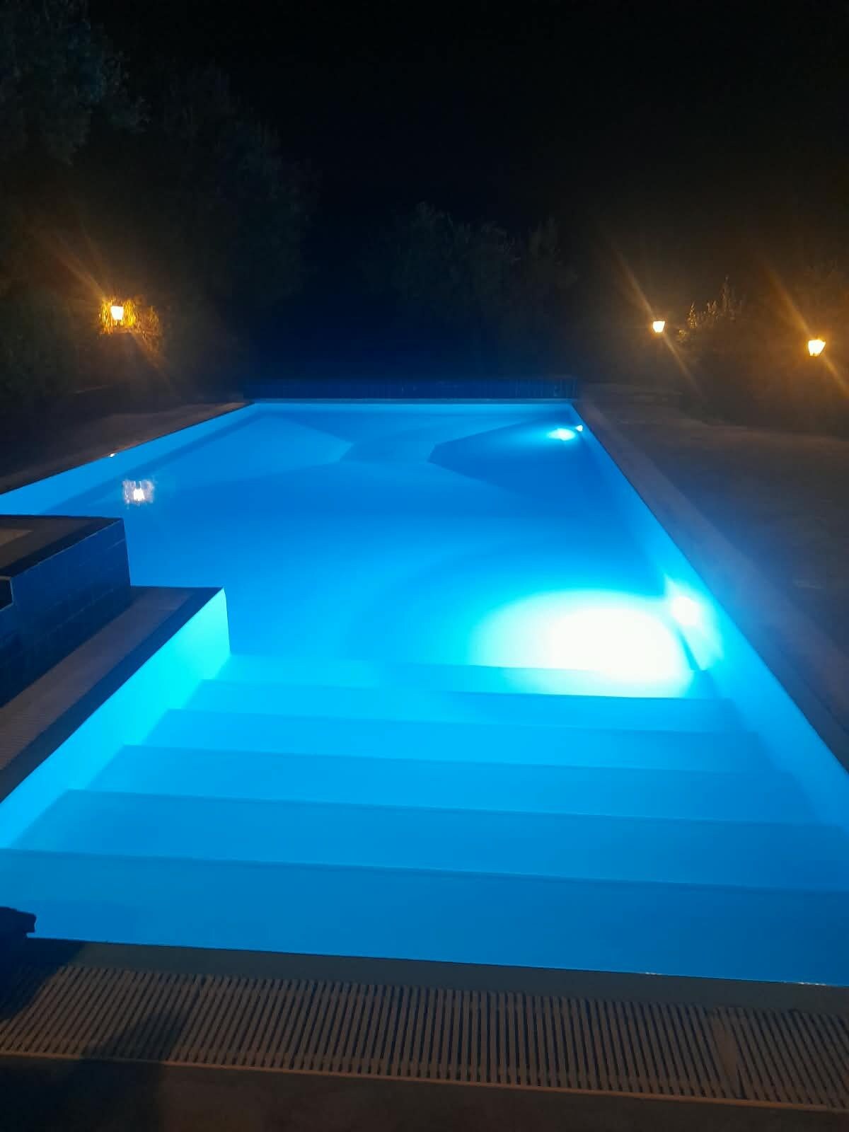 A rectangular swimming pool at night with steps leading into it, surrounded by walls with small lights.