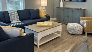 Een moderne woonkamer met een donkerblauwe bank, een witte salontafel, een houten voetenbankje en een aan de muur bevestigde televisie.