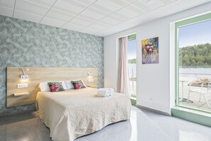 A modern bedroom with a large bed, a headboard with integrated lighting, a view of the outdoors through a large window, and a painting on the wall. - Les Begues Apartments (Begues)
