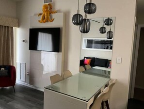 Een moderne woonkamer met een eettafel met glazen blad, beige stoelen, een rode fauteuil, een wandgemonteerde televisie en een grote spiegel met hanglampen.