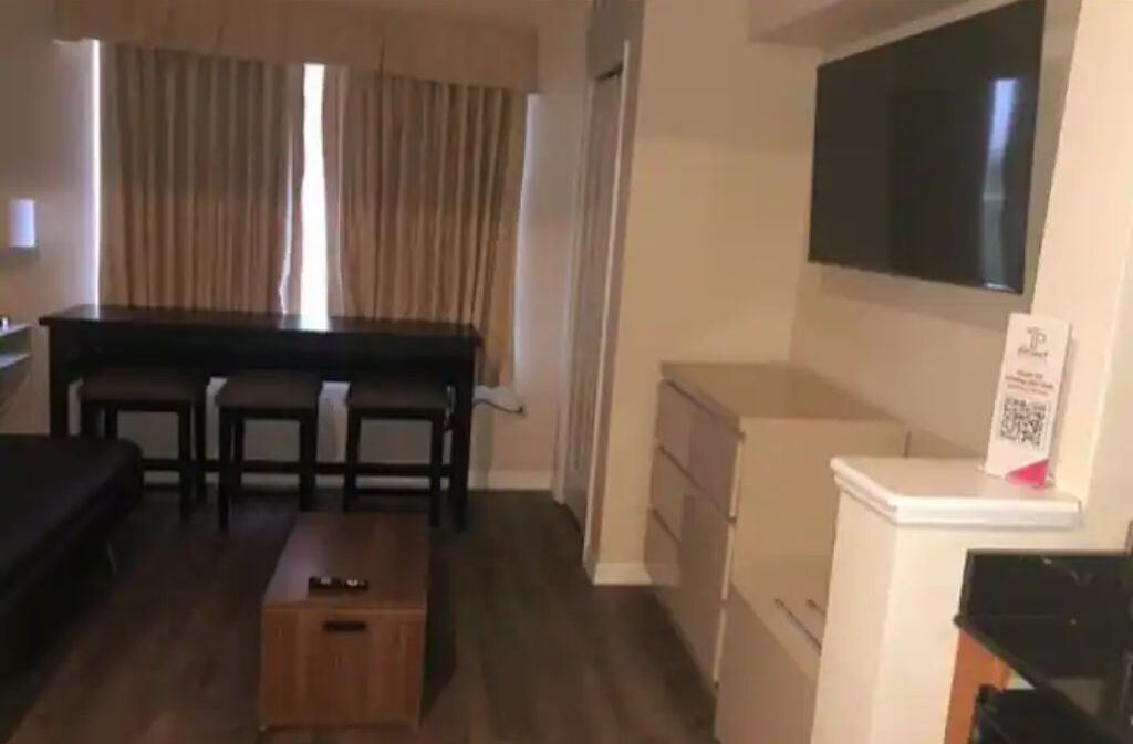 A compact living space with a sofa, a small wooden table, a TV mounted on the wall, and a kitchenette area.