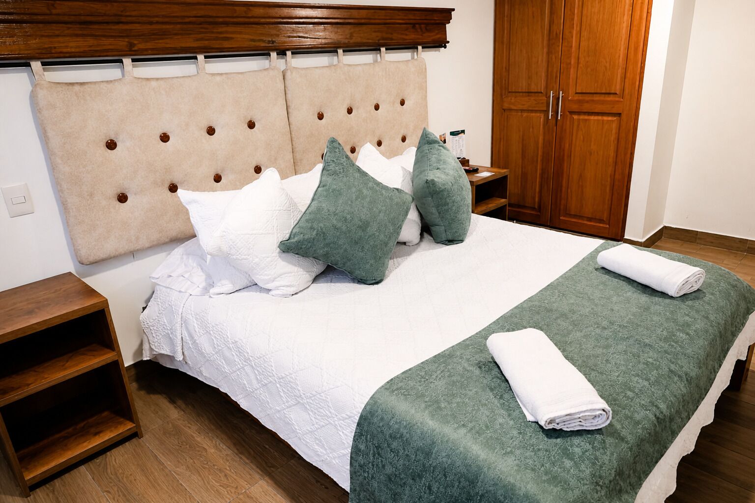A neatly made bed with a headboard, pillows, a green blanket, and a rolled towel.