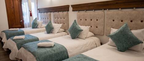 A hotel room with four beds, each with a tufted headboard, white bedding, and teal throw pillows.