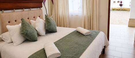 A neatly made bed with pillows and a rolled towel, a wooden headboard, and a view of the outside through a window with curtains.